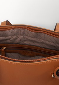 Innenseite einer tanfarbenen Ledertasche mit einem gesteppten, braunen Stofffutter. Verfügt über eine Reißverschlusstasche und goldfarbene Beschläge.