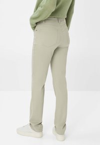 Persona che indossa pantaloni beige a gamba dritta e sneakers bianche, in piedi con le mani in tasca, su uno sfondo bianco semplice.