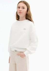 Bílá mikina se vzorem hvězdy a vyšitým textem "JSTE ÚŽASNÍ" na přední straně, doplněná světle béžovými kalhotami.