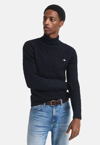 Pulover cu guler înalt, tricotat cu model în formă de cabluri, de culoare bleumarin, cu manșete și tiv din tricot cu nervuri. Prezintă un mic logo brodat pe piept. Purtați-l alături de blugi din denim de culoare bleumarin deschis.