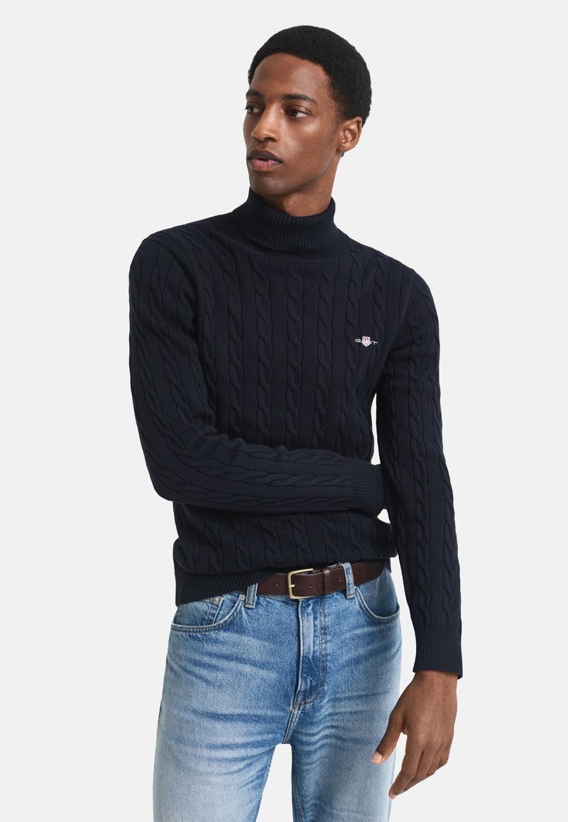 Pulover cu guler înalt, tricotat cu model în formă de cabluri, de culoare bleumarin, cu manșete și tiv din tricot cu nervuri. Prezintă un mic logo brodat pe piept. Purtați-l alături de blugi din denim de culoare bleumarin deschis.