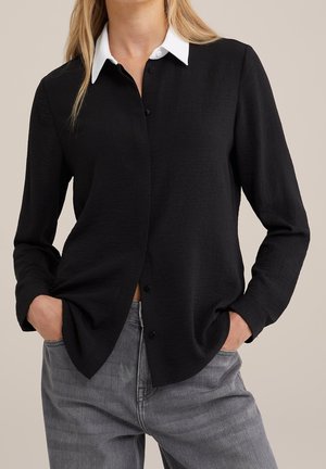 Vrouw die een zwart shirt met lange mouwen en een witte kraag draagt, en grijze spijkerbroek, handen in de zakken, tegen een effen achtergrond.