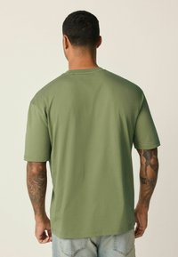 Oliivinvihreä puuvillainen t-paita, jossa on lyhyet hihat ja pyöreä pääntie, rento istuvuus ja sileä pinta. Takakuvassa näkyy yksinkertainen design.