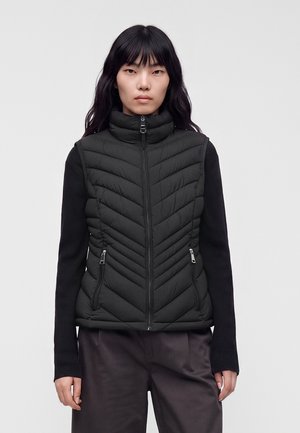 Frau mit langen schwarzen Haaren, die eine schwarze gesteppte Weste über einem schwarzen Langarmshirt und dunklen Hosen trägt, vor einem schlichten hellen Hintergrund stehend.