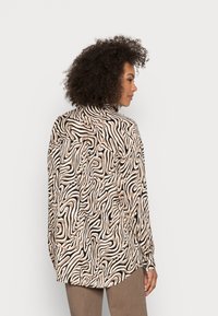 Blusa de manga longa em bege com padrão de ondas pretas, apresentando um corte descontraído e um colarinho clássico, vista pela parte de trás com uma bainha curva.