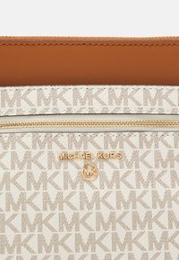 Pojemnik Michael Kors w kolorze kremowym z powtarzającym się wzorem logo MK. Posiada złoty zamek błyskawiczny oraz brązowy skórzany panel górny z wytłoczonym logo.