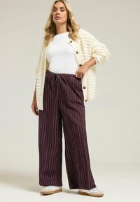 Cardigan en peluche crème avec des boutons marron, haut ajusté blanc et pantalon large bordeaux à rayures. Chaussures décontractées avec des tons blancs et beiges.