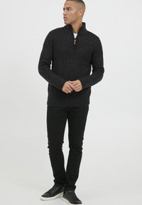 Suéter negro de punto con cuello alto y cremallera, que presenta un patrón texturizado, acompañado de pantalones negros y zapatos negros.