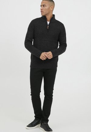 Schwarzer Strickpullover mit hohem Kragen und Reißverschluss, ausgestattet mit einem strukturierten Muster, kombiniert mit schwarzen Hosen und schwarzen Schuhen.
