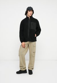 Schwarze Fleecejacke mit Reißverschluss, ausgestattet mit einer glänzenden schwarzen Taschenklappe. Kombiniert mit beigen Cargo-Hosen und schwarzen Sneakers.