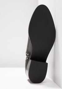Black leather shoe sole with block heel, studded leather upper, displayed against white background and wall corner.