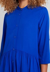 Robe bleue à manches longues avec un col boutonné, une partie inférieure froncée et une texture lisse. Présente une coupe décontractée et des coutures incurvées.