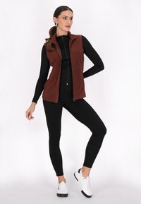 Vest de fleece maro cu fermoar frontal, asortat cu un top negru cu mânecă lungă și colanți negri, completat de adidași albi.