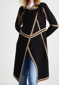 Schwarzer Strickmantel mit goldenen geometrischen Streifen, ausgestattet mit einem seitlichen Bindebandverschluss und einer eleganten, verlängerten Silhouette. Getragen über blauen Jeans.