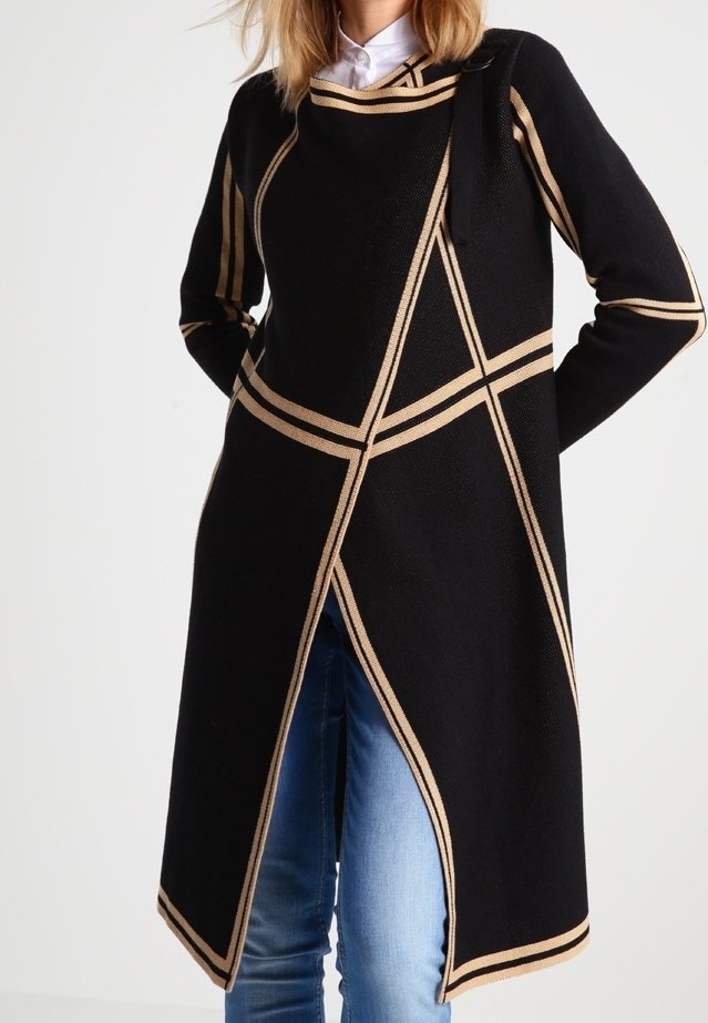 Schwarzer Strickmantel mit goldenen geometrischen Streifen, ausgestattet mit einem seitlichen Bindebandverschluss und einer eleganten, verlängerten Silhouette. Getragen über blauen Jeans.