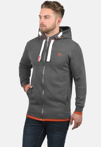 Sweat à capuche zippé gris avec une capuche doublée, des accents rouges et des cordons blancs. Dispose d'un logo sur la poitrine et de bords côtelés aux poignets et à l'ourlet.