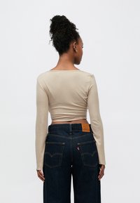 Bēša ilgtermiņa piedurkņu blūze ar kvadrātveida kakla izgriezumu, kombinēta ar tumšām džinsa biksēm ar izteiktām aizmugurējām kabatām un brūnā krāsā marķētu etiķeti.