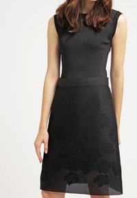 Černé šaty bez rukávů s žebrovaným živůtkem a květinovým krajkovým vzorem na sukni, s hladkým látkovým páskem v pase.