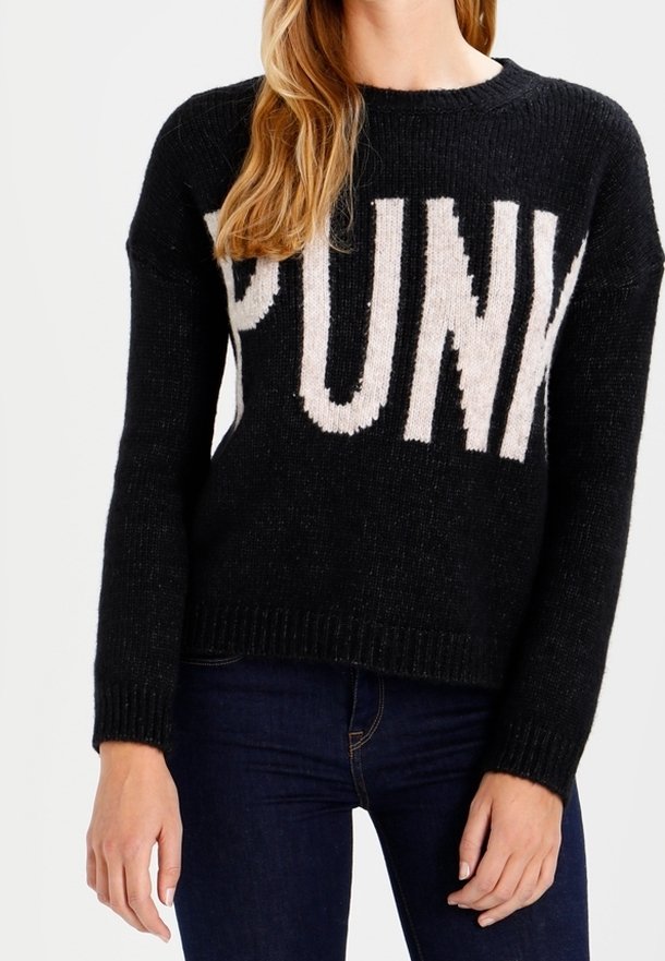 Femme portant un pull en maille noir avec de grandes lettres blanches formant le mot "PUNK" sur la poitrine et un jean bleu foncé.
