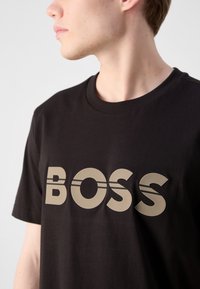 Melnas kokvilnas T-krekls ar lielu beigu "BOSS" logotipu un svītrainām akcentiem. Īsām piedurknēm un klasiskā apaļā kakla dizains.