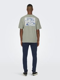 Groen T-shirt met korte mouwen, met een blauwe bergafbeelding op de achterkant en de tekst "Wild Spirit", in combinatie met donkerblauwe jeans en zwarte sneakers.