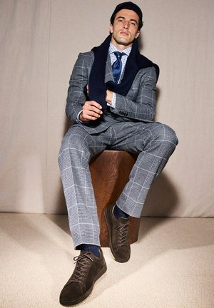 Costume à carreaux gris avec une coupe ajustée, associé à une écharpe navy et des baskets en daim marron foncé. Tabouret en bois en arrière-plan.
