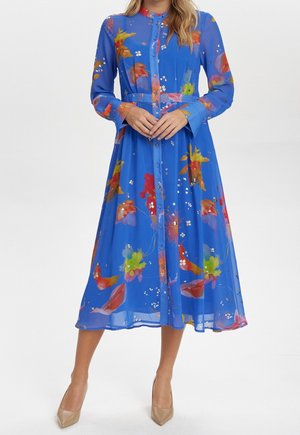 Kobieta w niebieskiej sukience midi z długim rękawem i kolorowym kwiatowym wzorem, z przodu zapinanej na guziki, w beżowych szpilkach, stojąca na białym tle.