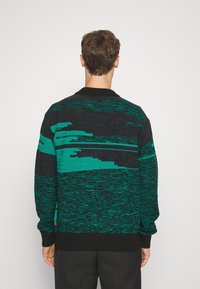 Pull en tricot noir et vert, présentant des motifs horizontaux abstraits. Tissu texturé avec poignets et col côtelés.