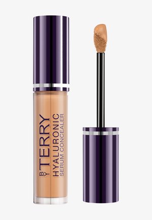 Tube de correcteur avec un bouchon violet foncé et un applicateur en baguette montrant la crème correctrice beige sur une pointe d’éponge inclinée.