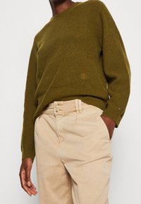 Osoba v olivově zeleném žebrovaném svetru a béžových kalhotách s vysokým pasem a dvojitým zapínáním, s jednou rukou v kapse.