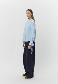 Hellblaues Hemd mit Kragen, langen Ärmeln und gebundenen Öffnungen, kombiniert mit dunklen, weiten Hosen. Einfaches Design, glatte Textur.