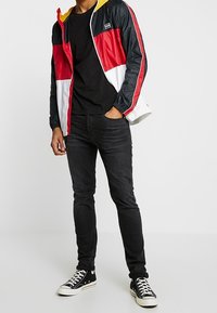 Mężczyzna ubrany w kurtkę z blokami kolorów w czerni, czerwieni, bieli i żółci, czarny T-shirt, czarne wąskie jeansy oraz czarne wysokie trampki z białymi sznurowadłami.