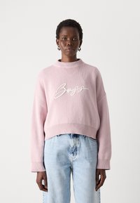 Lyserød strikket sweater med høj hals og sænkede skuldre, prydet med en hvid broderet "Boss"-logo på forsiden. Bæres med blå jeans.