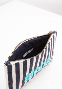 Pochette en tissu rayé bleu marine et crème. Elle possède une fermeture éclair, une doublure bleu marine et une broderie turquoise "aloha" à l'avant.