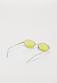Gafas de sol con tinte amarillo, lentes ovalados, montura de metal plateado y varillas delgadas. Incluyen almohadillas para la nariz y detalles de acento en las patillas.