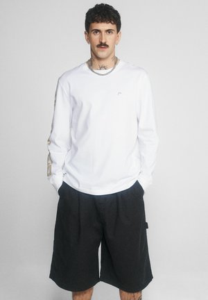 Mężczyzna z kręconymi włosami i wąsami, ubrany w białą koszulę na długi rękaw i czarne krótkie spodenki do kolan, stojący z rękami w kieszeniach.