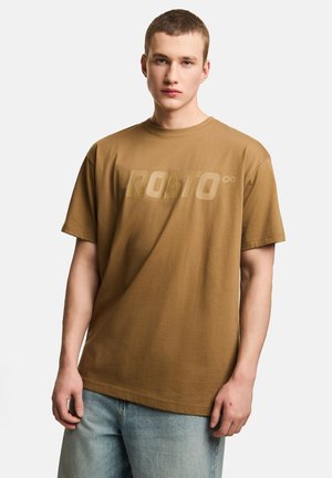 Młody mężczyzna o krótkich włosach, ubrany w brązowy t-shirt PROSTO i jasnoniebieskie dżinsy, stojący na jednolitym białym tle.