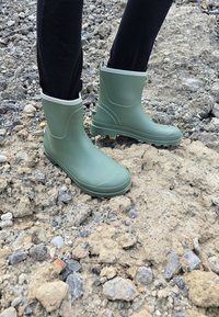 Bottes en caoutchouc vertes imperméables avec une semelle texturée, hauteur cheville, et une bordure blanche contrastante, posées sur une surface en gravier.
