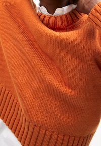 Gros plan d'une personne portant un pull orange texturé en maille par-dessus une chemise blanche à col, ajustant le pull près du cou.