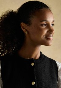 Must must vest, mille ümarate kuldsete nööpidega, kantud valge pluusi kohal. Juuksed on lahtiste lokkidega, rõhutades kuldseid kirurgi kõrvarõngaid.