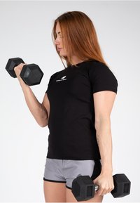 Melnas īsām piedurknēm krekls ar logo, pelēki šorti, turot melnus sešstūrainus hanteles, uz kurām norādīts 6 kg, rādīts sānu profila skats.