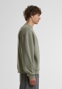 Oliven grøn sweatshirt med rund hals og ribbede manchetter, som har en afslappet pasform og blød tekstur. Modellen vises fra siden.