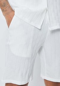 Shorts blancs dotés d'un motif texturé avec un design géométrique, des poches latérales et un ourlet droit. Le tissu semble léger et décontracté.
