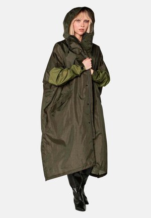 Vrouw draagt een lange, oversized olijfgroene regenponcho met capuchon en zwarte laarzen, staand tegen een effen lichte achtergrond.