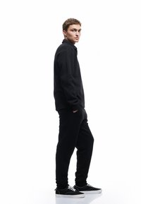 Sort sweatshirt med høj krave, parret med sorte sweatpants. Modellen har sorte sneakers med hvide såler på og står foran en hvid baggrund.