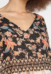 Vêtement noir à col en V avec des motifs floraux et cachemire complexes en nuances d'orange, de bleu et de beige, porté par une personne aux cheveux bouclés.