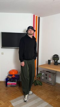 Črna jopa s kapuco, široke zelene hlače in sivi športni copati. V ozadju televizor, oranžna svetilka in lončnica na lesenem tleh.