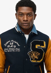 Chaqueta tipo varsity de lana azul marino con mangas de cuero naranja, cuello de denin, texto bordado y un gráfico de tigre. Cierre a presión en la parte frontal.