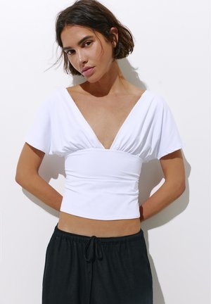 Une femme porte un haut blanc court à col en V et à manches courtes, ainsi qu’un pantalon noir à cordon de serrage, devant un mur uni.