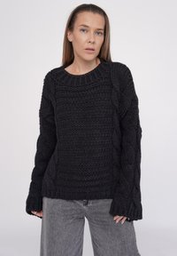 Schwarzer Strickpullover mit strukturiertem Zopfmuster, rundem Ausschnitt und langen, weiten Ärmeln. Kombiniert mit hellgrauen Jeans.
